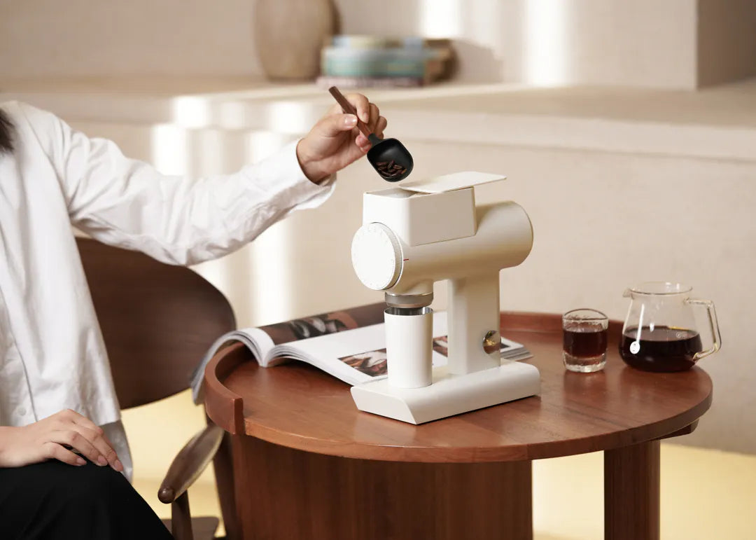 Person using TIMEMORE Sculptor 064s grinder on a wooden table with coffee glass and magazine.